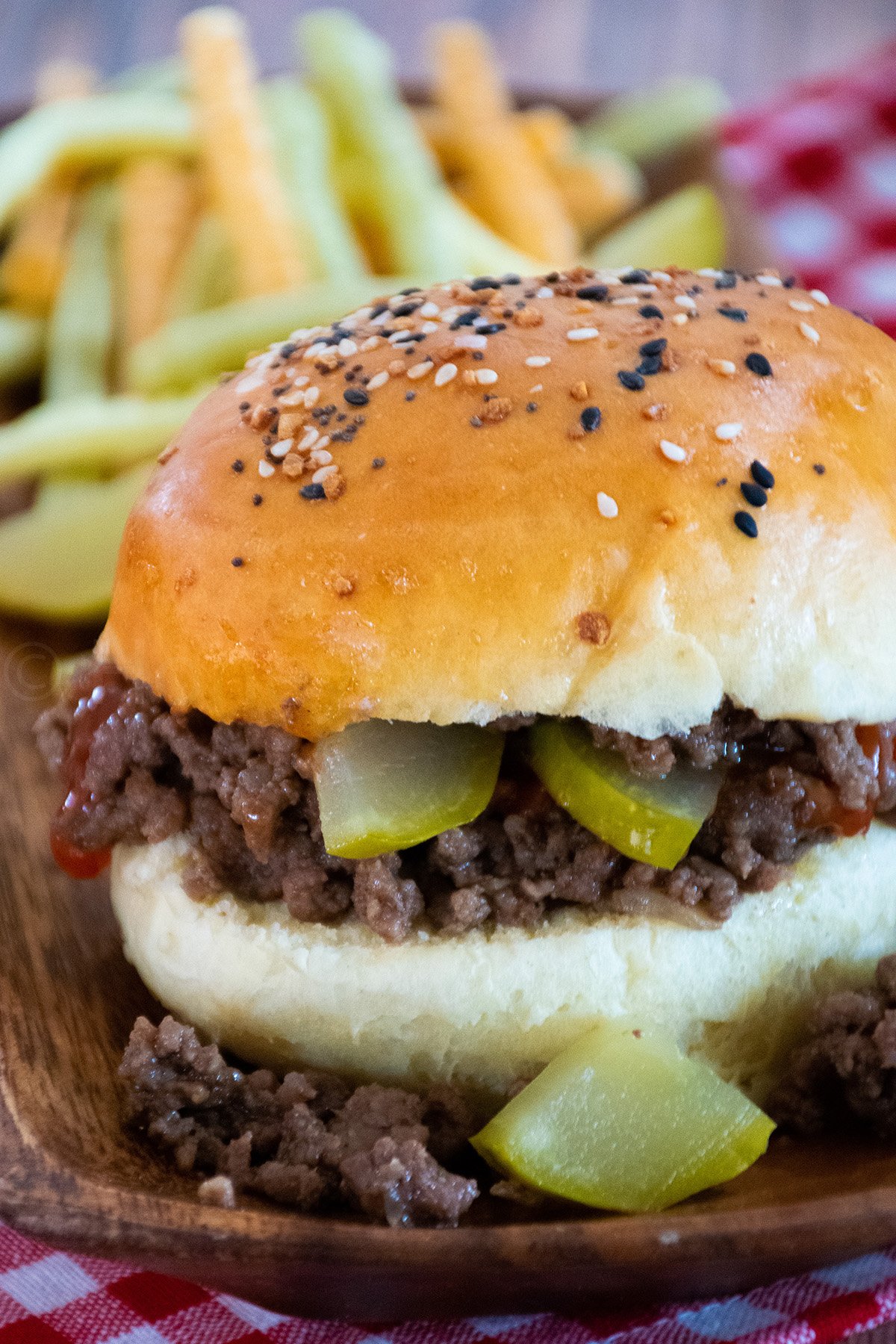 Loose meat sandwich served on a homemade bun topped with everything seasoning, filled with ground beef, dill pickles, and ketchup, with veggie fries in the background.