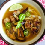 A bowl of spicy veal meatball pho soup with mushrooms, noodles, lime, and herbs.