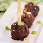 Close-up of glazed Mongolian meatballs on a white rectangular plate, garnished with sesame seeds and green onions, with wooden picks inserted for serving.