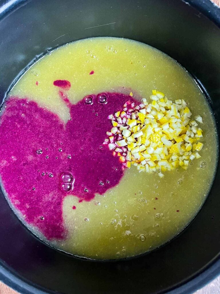 A pot showing a mixture of muscadine grape juice, bright purple skin puree, and finely chopped yellow lemon peel before stirring.