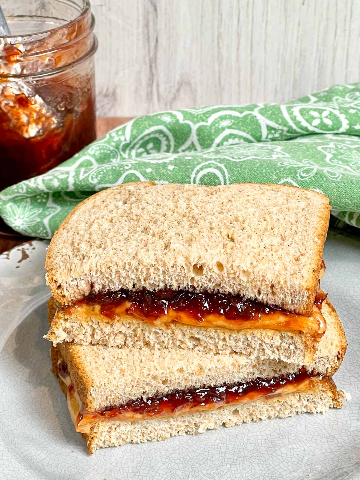 A classic peanut butter and. muscadine jam sandwich made with soft whole wheat bread, cut in half and stacked on a plate. In the background, a jar of muscadine jam with a spoon inside sits next to a patterned green cloth.