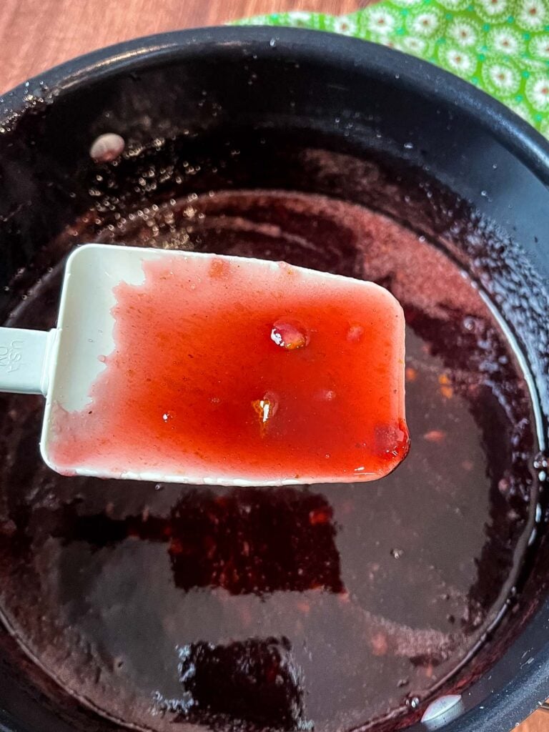 A close up of a white spatula coated in glossy red muscadine jam held over a dark pot.