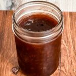 Jar filled with dark, glossy muscadine jam sitting on a wooden surface.
