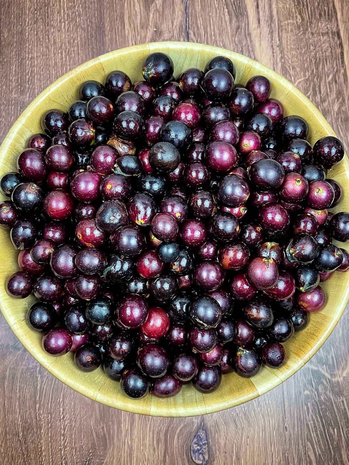 A wooden bowl filled to the brim with freshly washed, dark purple muscadine grapes on a rustic wooden surface.