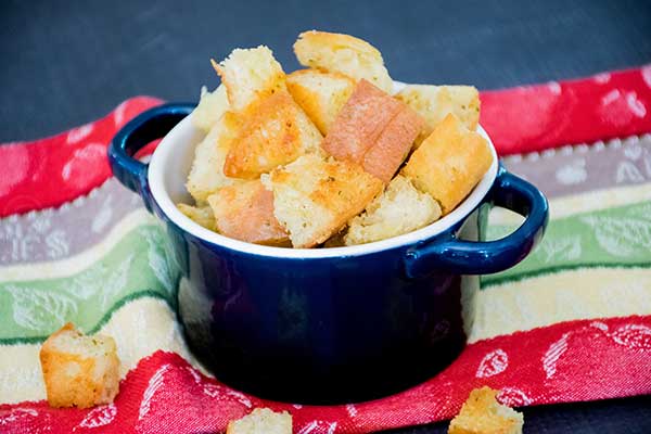 A small navy blue bowl filled with golden brown homemade croutons sits on a colorful kitchen towel with red, green, and cream stripes. A few croutons are scattered around the bowl.