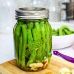 Close up of a glass jar filled with fresh green beans, garlic cloves, dill, mustard seeds, and red pepper flakes, sealed with a silver lid, sitting on a wooden cutting board.