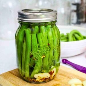 Close up of a glass jar filled with fresh green beans, garlic cloves, dill, mustard seeds, and red pepper flakes, sealed with a silver lid, sitting on a wooden cutting board.