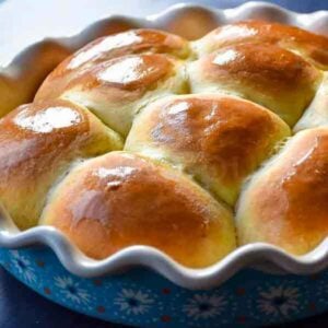 Freshly baked dinner rolls with a glossy, golden-brown top, nestled together in a decorative blue scalloped baking dish.