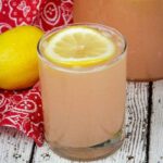 A glass of pink rhubarb lemonade garnished with a lemon slice, set on a rustic white wood surface beside a fresh lemon, a red patterned cloth, and a pitcher of lemonade in the background.