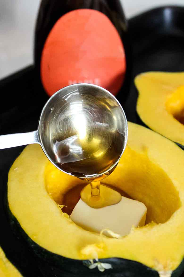 A close up of maple syrup being poured from a metal measuring spoon into a halved raw acorn squash with a pat of butter in the center.