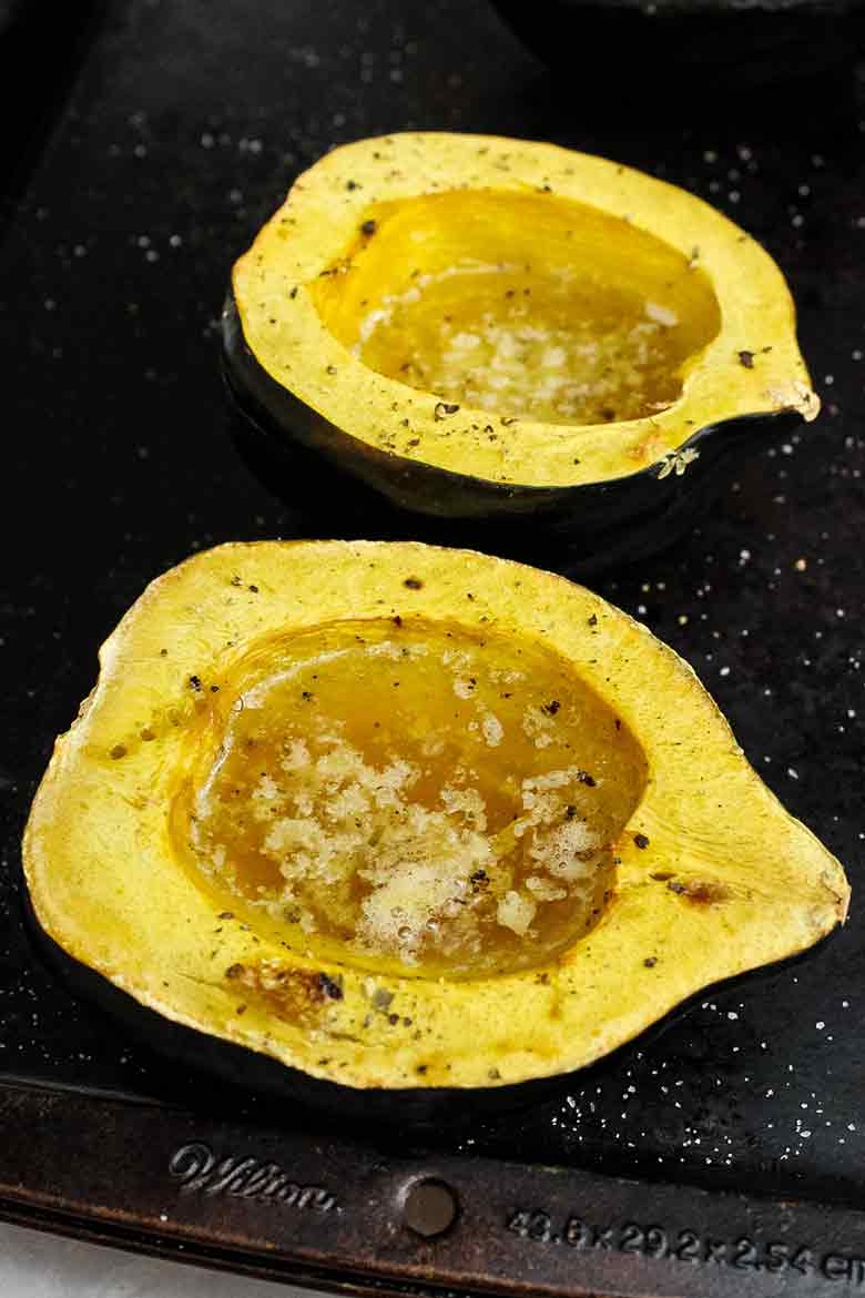 Two roasted acorn squash halves on a baking sheet, their centers filled with bubbling melted butter, syrup, and salt and pepper.
