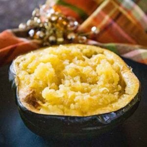 A halved roasted acorh squash filled with tender, mashed golden squash, served on a dark plate with a plaid cloth and beaded napkin ring in the background.