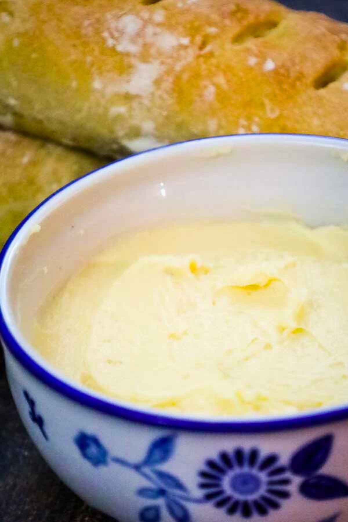 Close up of a bowl filled with creamy salted maple butter, with a loaf of crusty bread in the background.