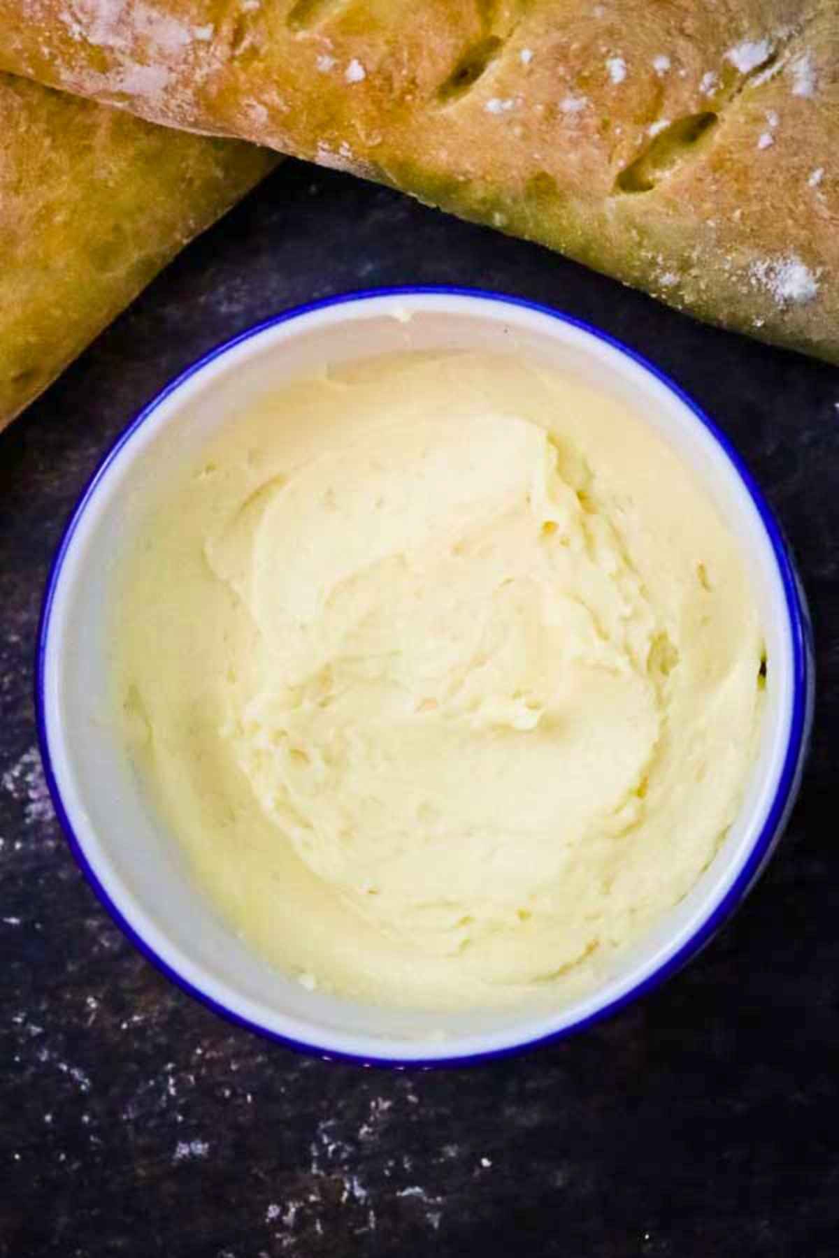 Overhead view of a bowl of salted maple butter on a dark countertop surrounded by slices of rustic bread.