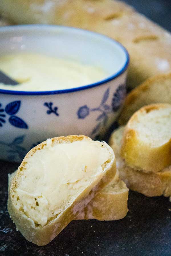 A slice of crusty bread spread with salted maple butter, with a decorative bowl of butter in the background.