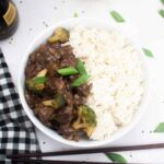 Top down view of a bowl of slow cooker beef teriyaki served with white rice and green onion garnish, with a black and white checked napkin and scattered green onion pieces around.