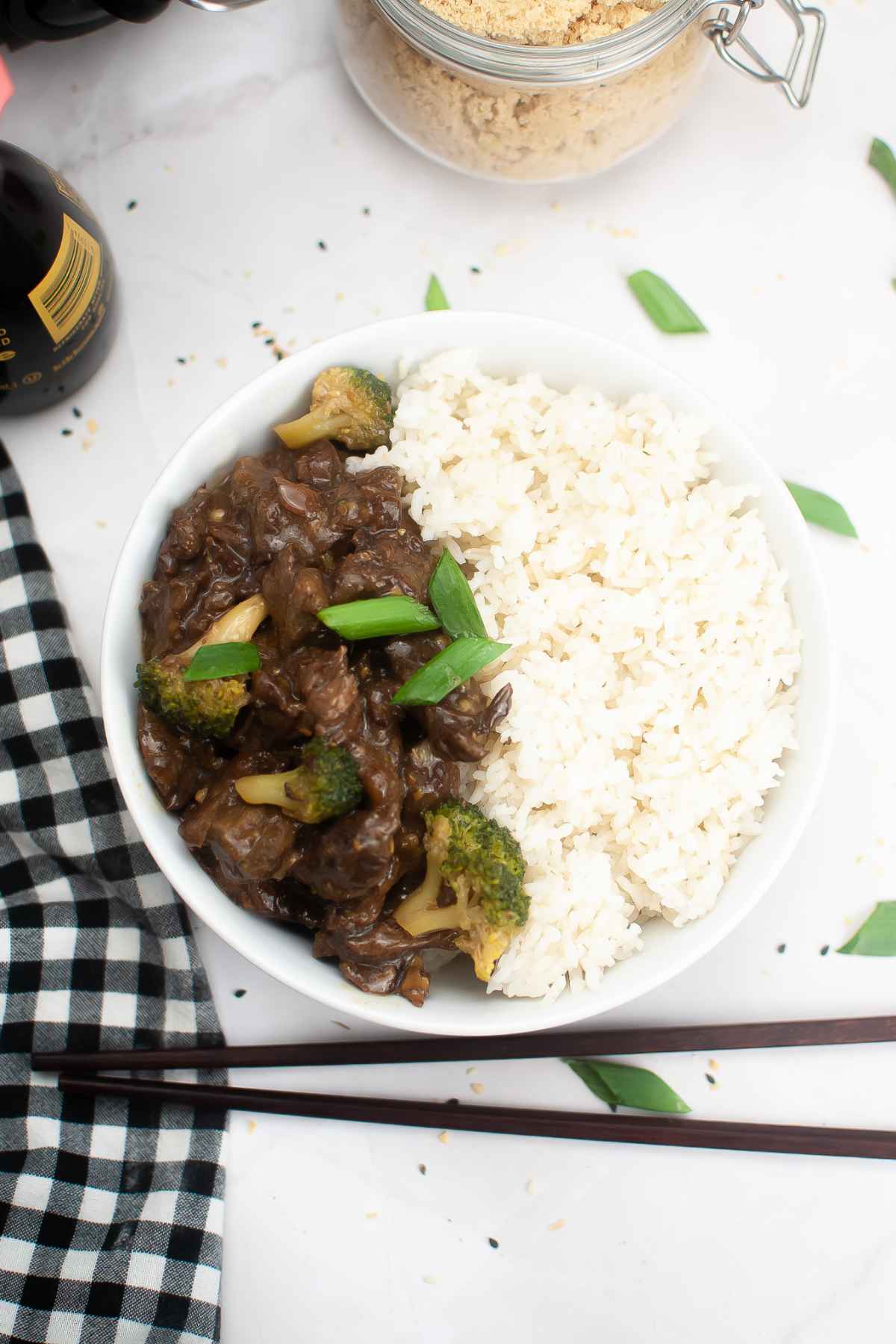Top down view of a bowl of slow cooker beef teriyaki served with white rice and green onion garnish, with a black and white checked napkin and scattered green onion pieces around.