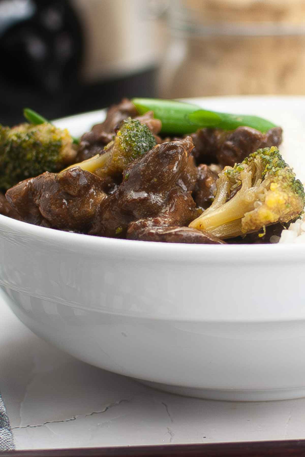 Close up side view of slow cooker beef teriyaki and broccoli in a white bowl with a rich, glossy sauce and visible green onion slices.