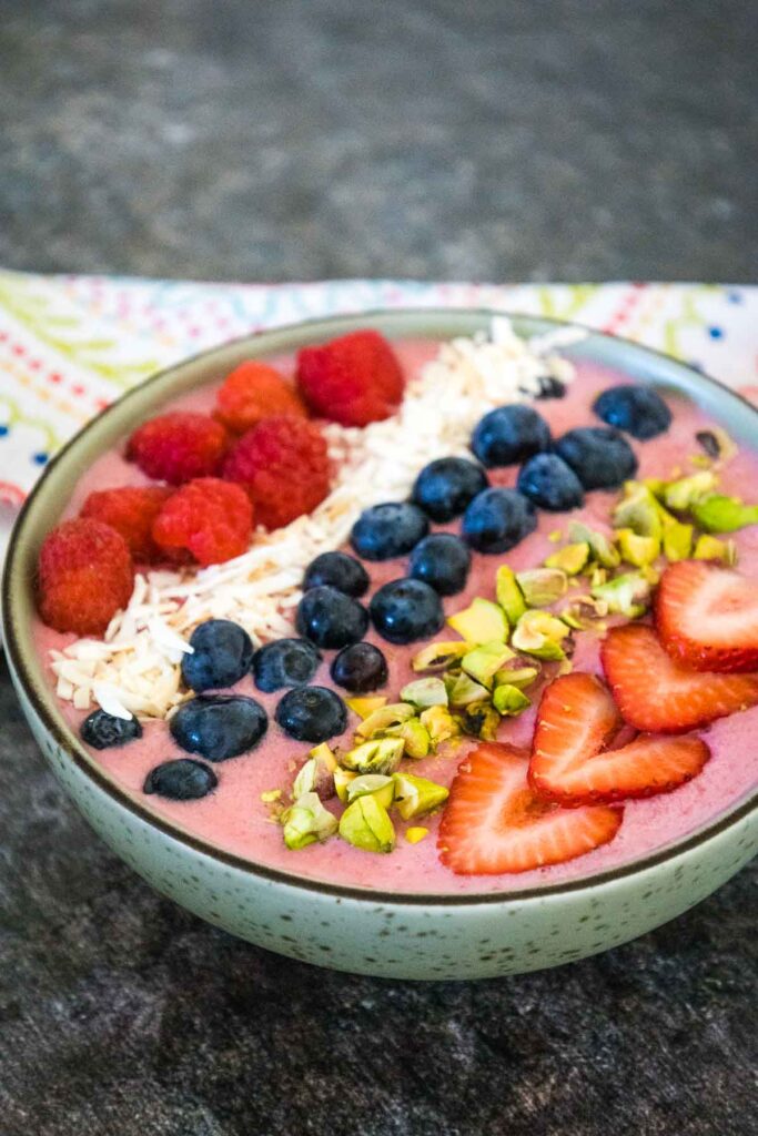 A vibrant smoothie bowl with a pink berry base, garnished with raspberries, blueberries, sliced strawberries, chopped pistachios, and shredded coconut.