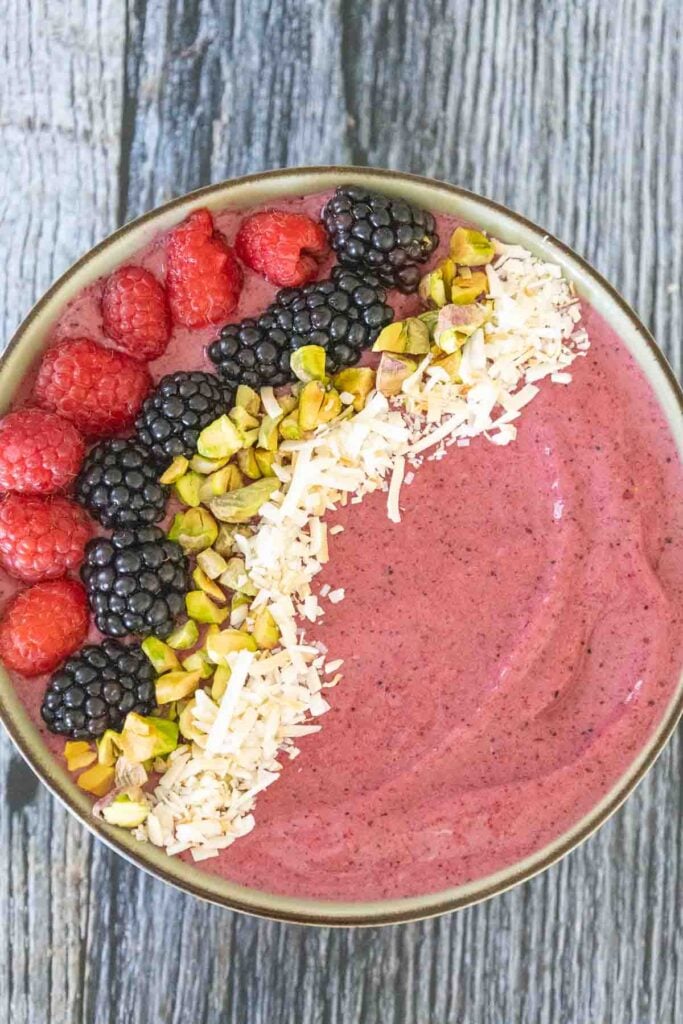 Overhead view of a pink berry smoothie bowl topped with neat rows of raspberries, blackberries, chopped pistachios, and shredded coconut on a wooden surface.