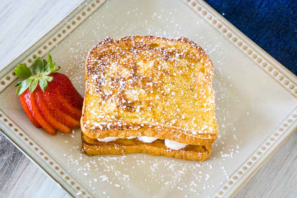 Top down view of french toast sandwich on a square tan plate, sprinkled with powdered sugar and served with a fan of sliced fresh strawberries.