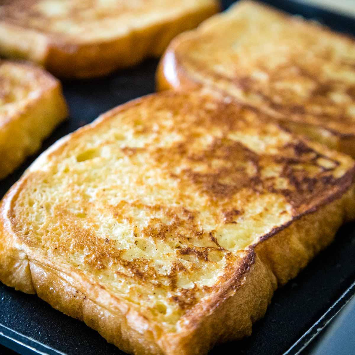 Several slices of golden french toast cooking on a flat griddle with crisp browned surfaces indicating they are almost ready to be stuffed.