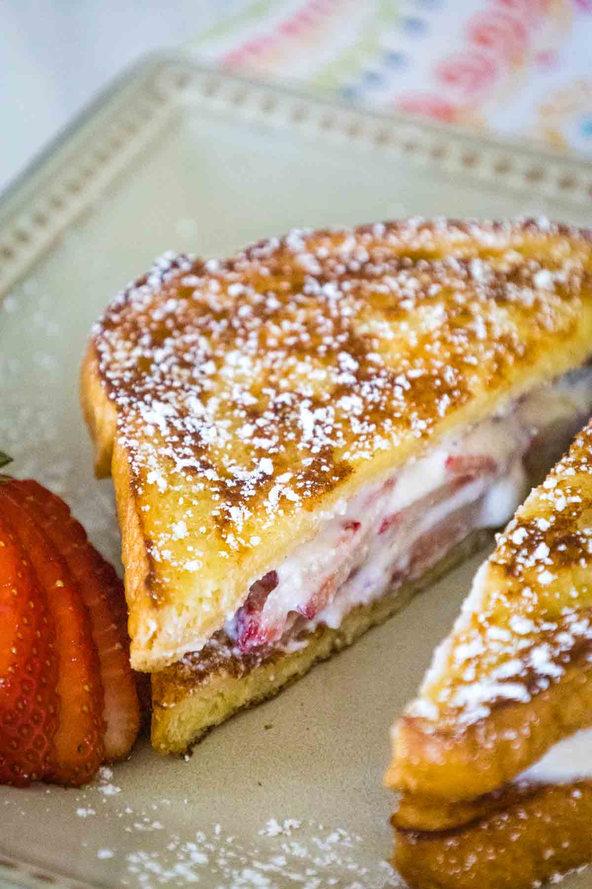 A close-up of a halved strawberry stuffed french toast sandwich showing a creamy filling with sliced strawberries, dusted with powdered sugar and served with fresh sliced strawberries on the side.