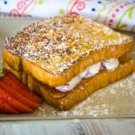 A golden brown stuffed french toast sandwich stacked with creamy filling peeking between the slices, dusted with powdered sugar and served with fresh strawberry slices on a tan plate.