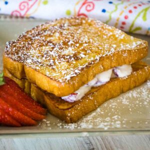 A golden brown stuffed french toast sandwich stacked with creamy filling peeking between the slices, dusted with powdered sugar and served with fresh strawberry slices on a tan plate.