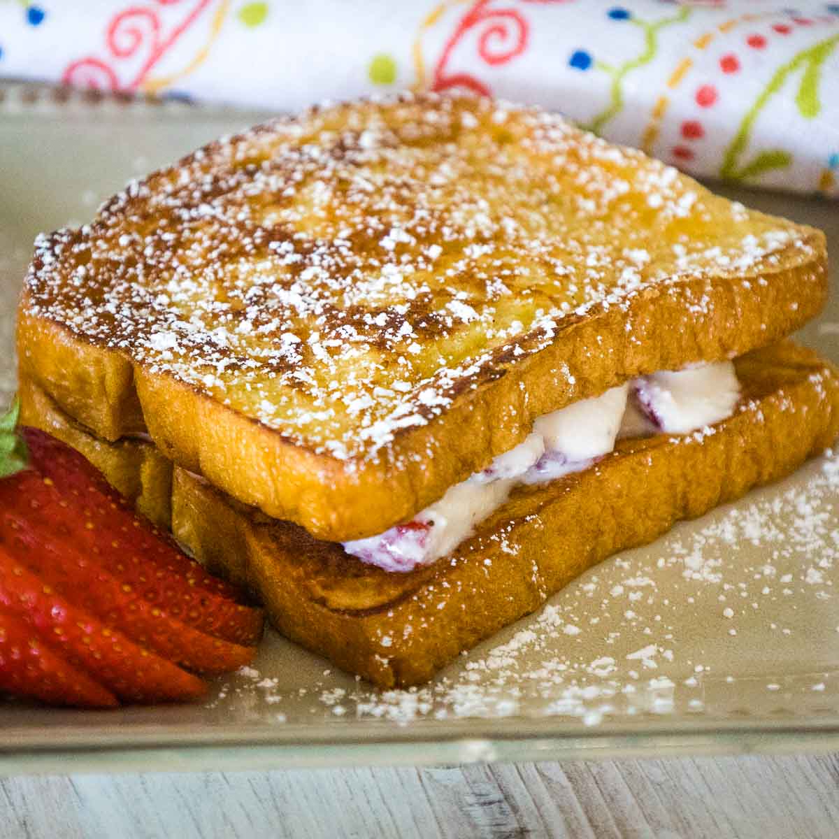 A golden brown stuffed french toast sandwich stacked with creamy filling peeking between the slices, dusted with powdered sugar and served with fresh strawberry slices on a tan plate.