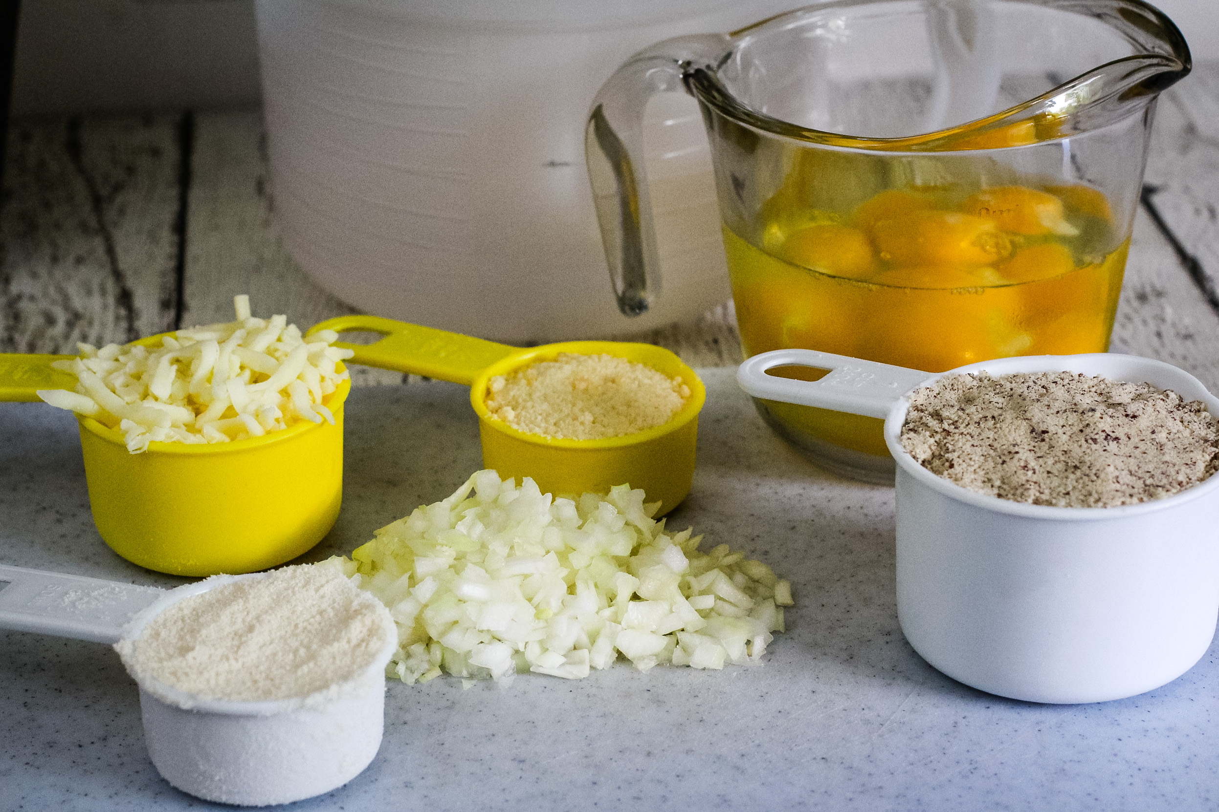 Overhead shot of measured ingredients on a counter, including chopped onion, shredded cheese, eggs, almond flour, and grated parmesan.