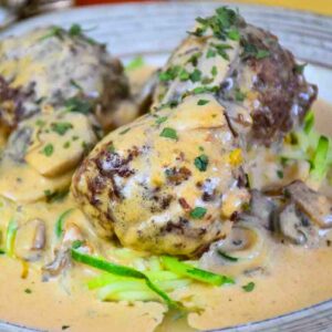 Up close image of three venison meatballs topped with creamy Alfredo mushroom sauce, served over a bed of zucchini noodles on a ceramic plate.