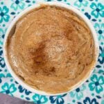 Overhead view of a white bowl filled with whipped cinnamon honey butter, topped with a light dusting of cinnamon, set on a teal and blue floral napkin.
