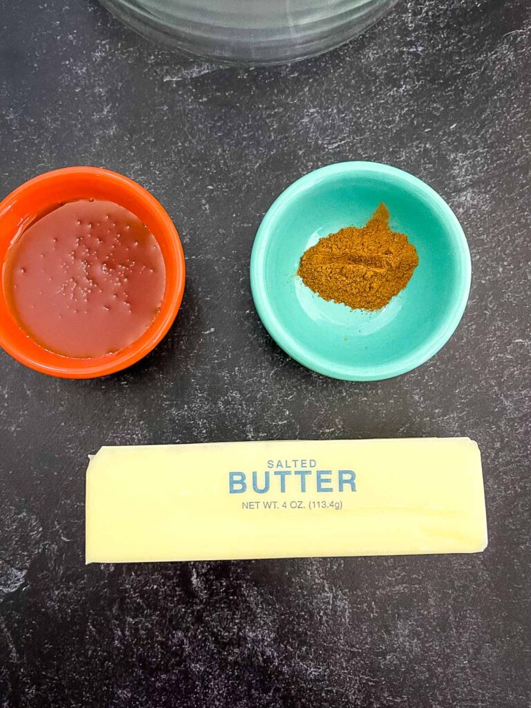 Flat lay of ingredients including a stick of salted butter, a small bowl of honey and a small bowl of ground cinnamon on a dark countertop.