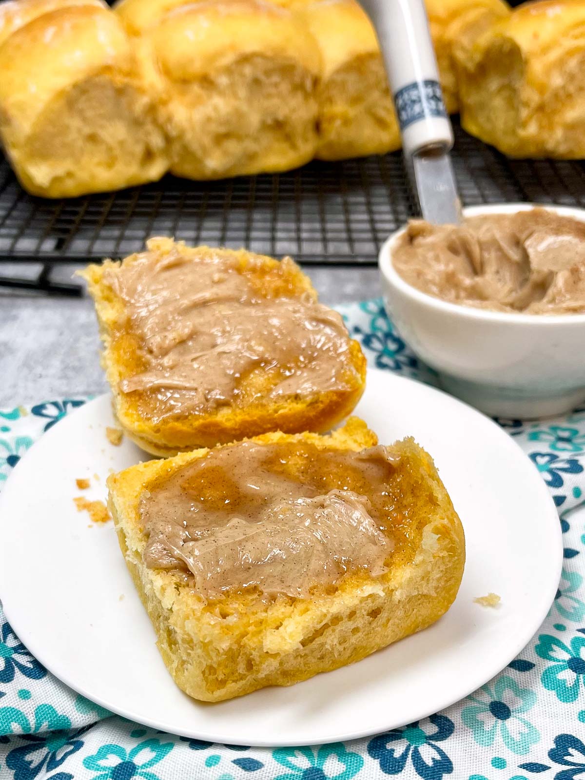 Fresh sweet potato rolls spread with whipped cinnamon honey butter on a white plate with a bowl of the butter and a tray of rolls in the background.