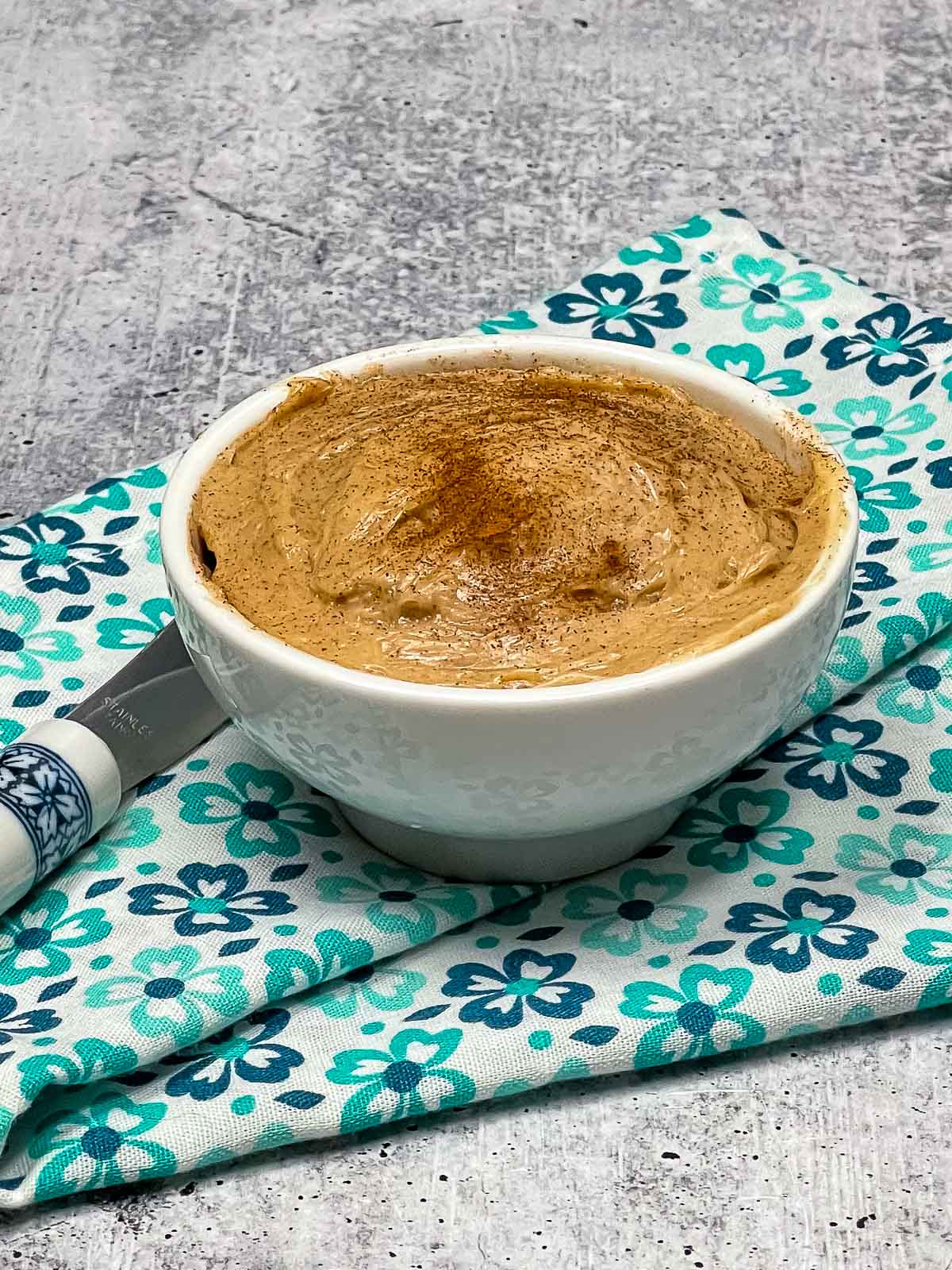 Small white bowl filled with whipped cinnamon honey butter, lightly dusted with cinnamon on top, set on a folded floral napkin with a butter knife beside it.