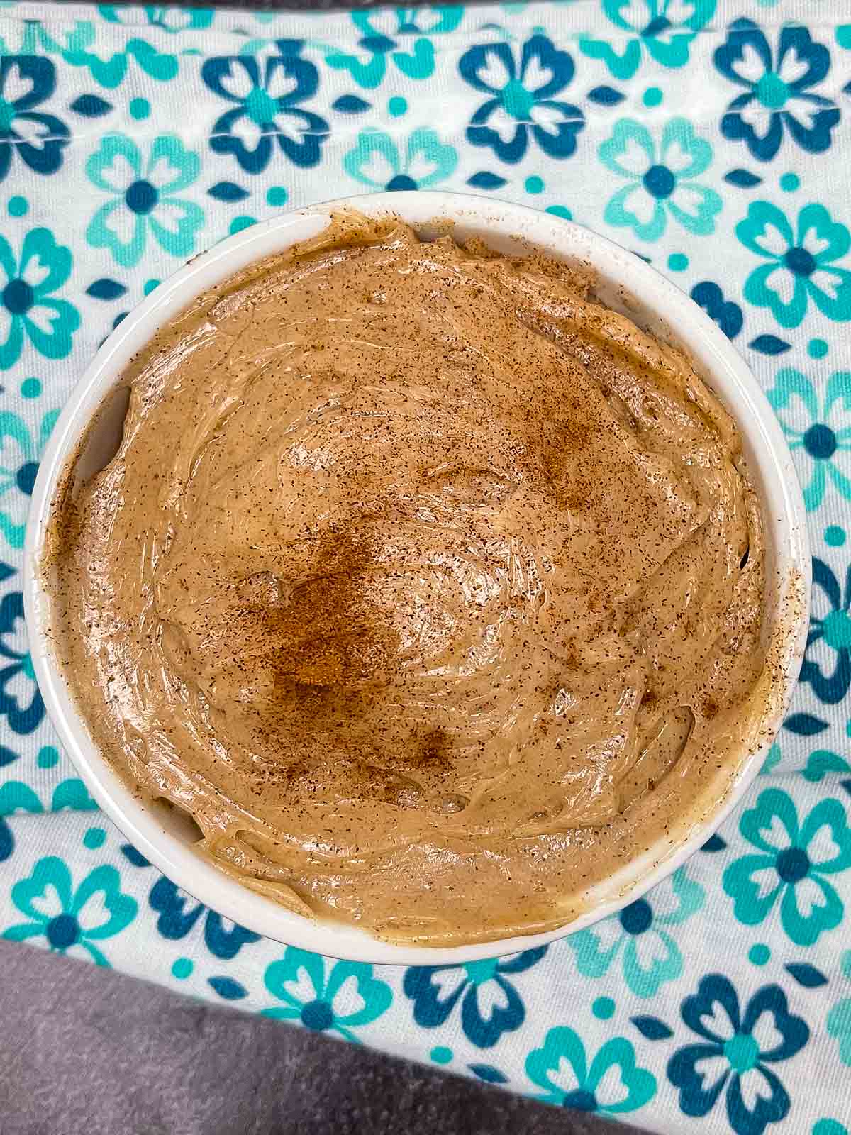 Overhead image of a small white bowl filled with whipped cinnamon honey butter topped with a dusting of cinnamon. The bowl is set on a teal and blue floral napkin.