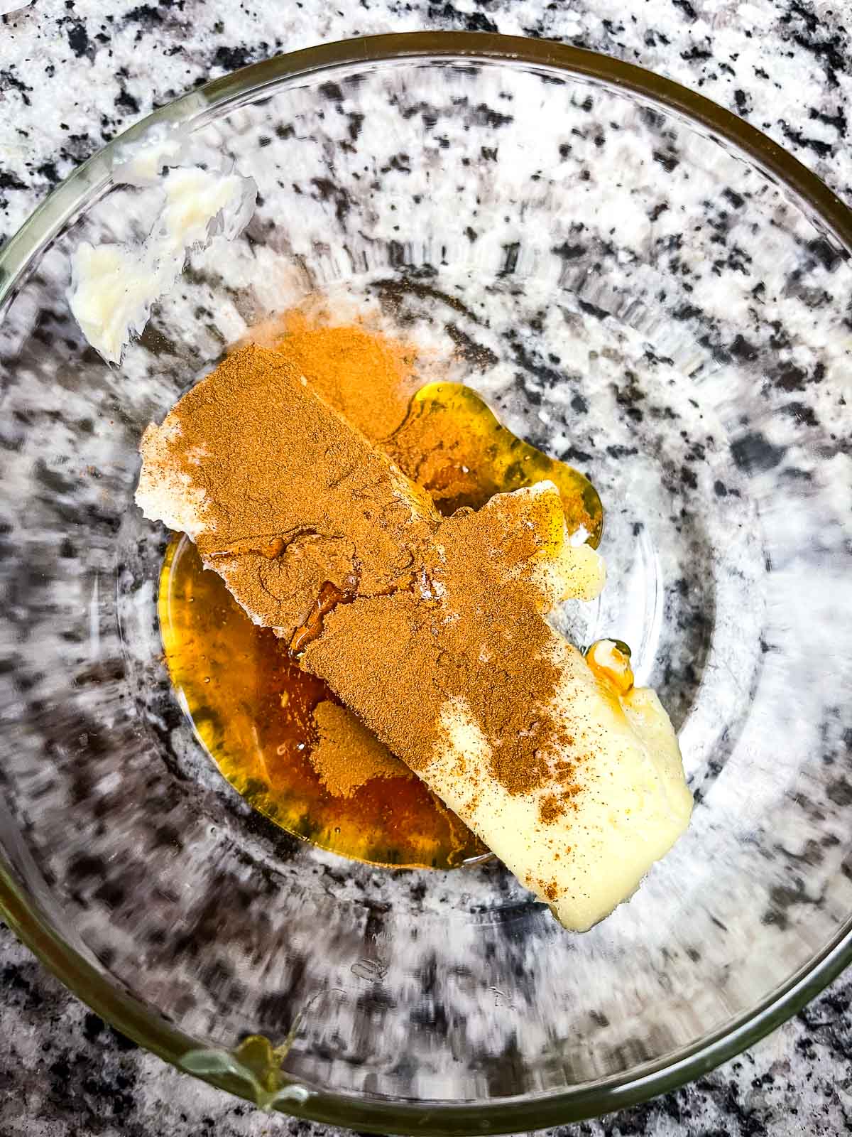 Overhead view of a glass mixing bowl containing a stick of butter, ground cinnamon, and honey, ready to be blended.