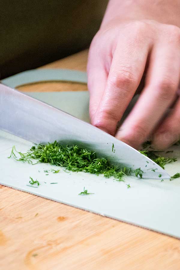 A close up of a hand using a chef's knife to finely chop fresh dill on a light cutting board, with a wooden countertop beneath.