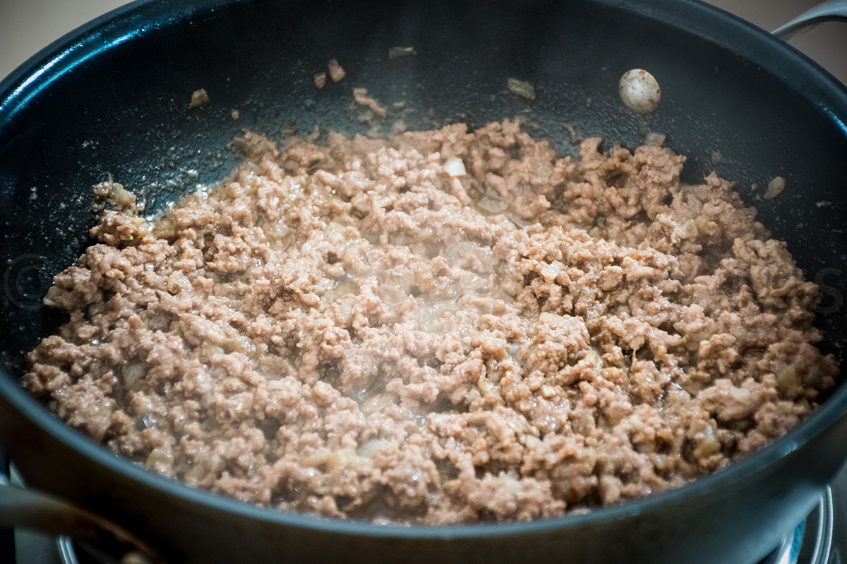 Ground beef and onion miixture cooking in a black skillet, steaming and browning.
