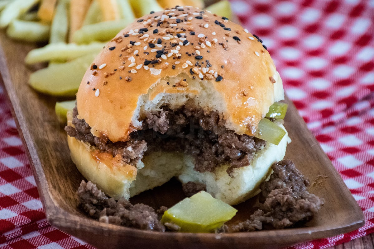 Loose meat sandwich with a bite taken out, showing seasoned ground beef and pickles on a soft homemade bun served with veggie fries on a red gingham tablecloth.