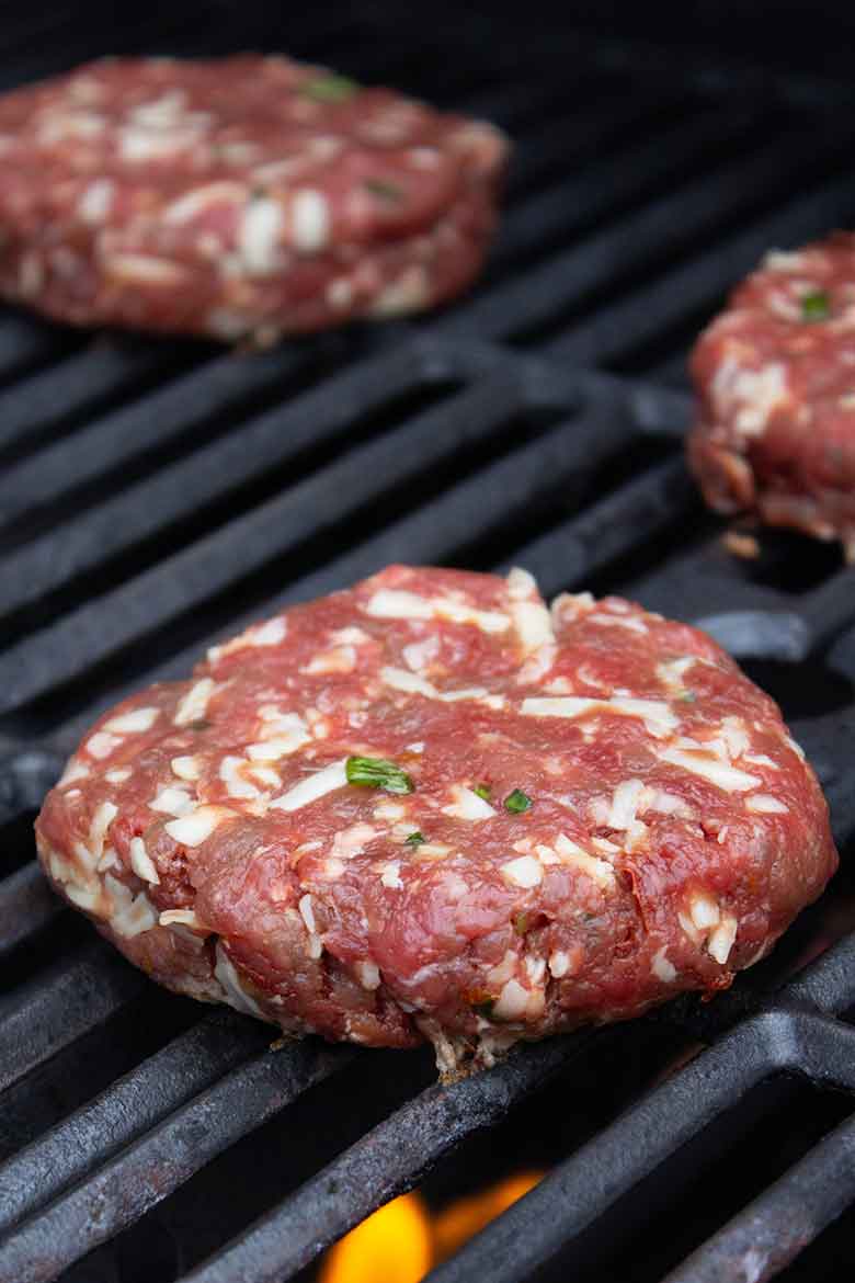 Raw venison burger patties on a grill with visible bits of shredded cheese and herbs in the meat mixture, ready for cooking.