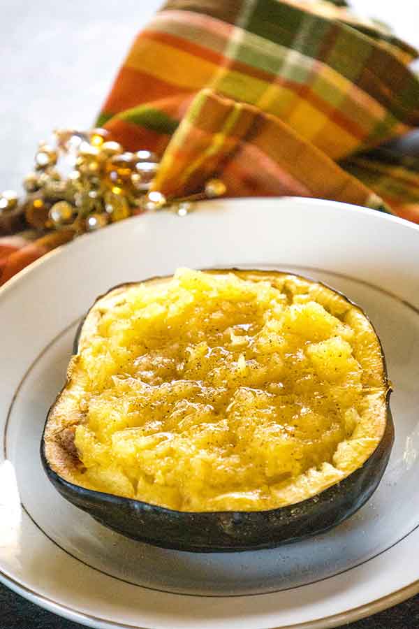 A halved roasted acorh squash filled with tender, mashed golden squash, served on a dark plate with a plaid cloth and beaded napkin ring in the background.