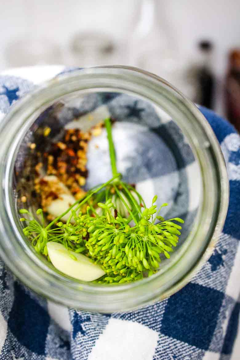 Top down view of a glass jar with garlic cloves, fresh dill heads, crushed red pepper, and mustard seeds layered at the bottom, resting on a blue and white checkered towel.