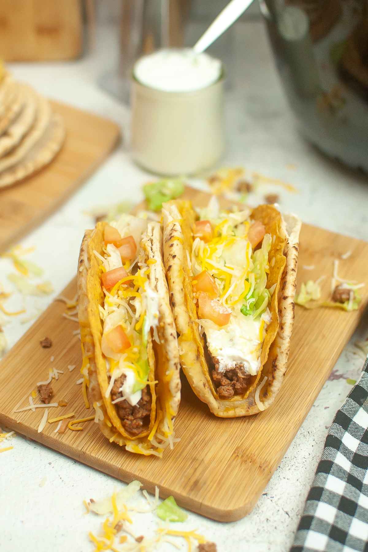 Two cheesy gordita crunch tacos filled with seasoned meat, lettuce, shredded cheese, diced tomatoes, and sour cream on a wooden board.