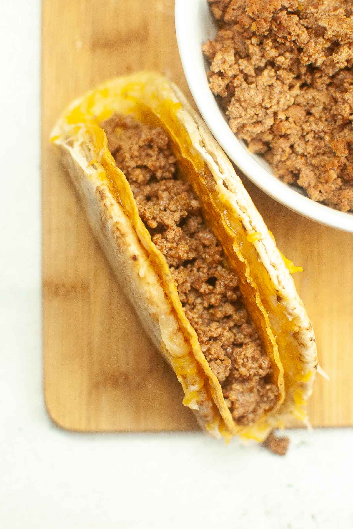 A cheesy gordita crunch being filled with seasoned ground beef, with a bowl of meat placed next to it on a wooden surface.