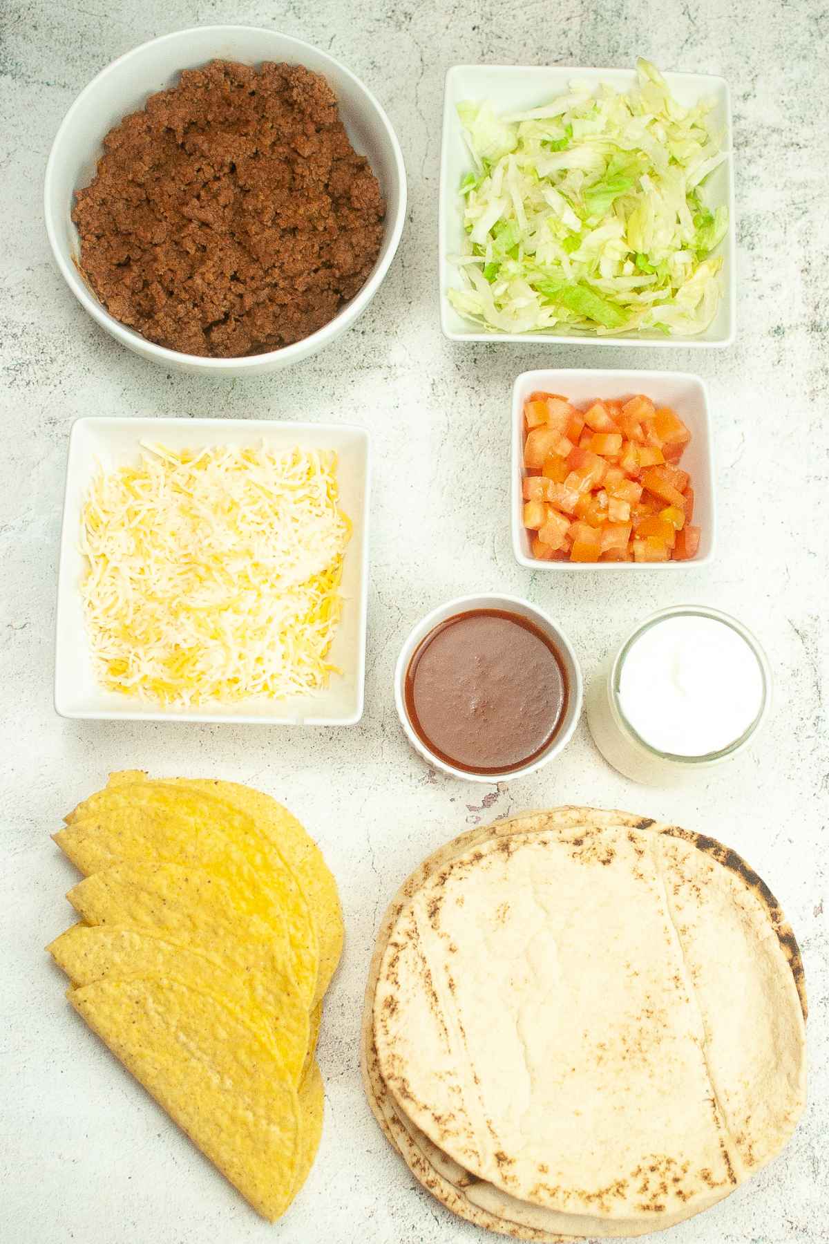 Overhead view of taco ingredients in small bowls and on the counter: seasoned ground meat, shredded lettuce, diced tomatoes, shredded cheese, taco sauce, sour cream, hard taco shells, and flatbread.