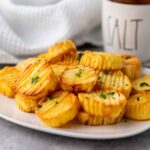 A plate of golden brown accordion potatoes garnished with chopped parsley served on a white plate with a soft gray background.