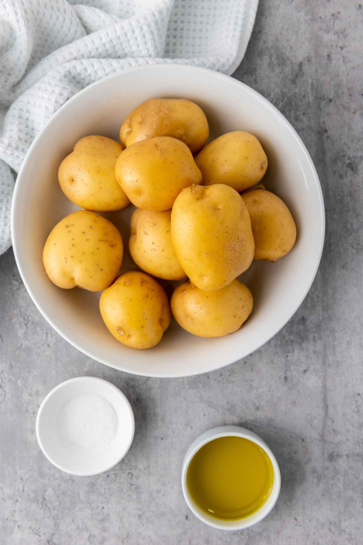 A bowl of raw yellow potatoes next to small dishes of salt and olive oil, set on a gray surface with a white towel.