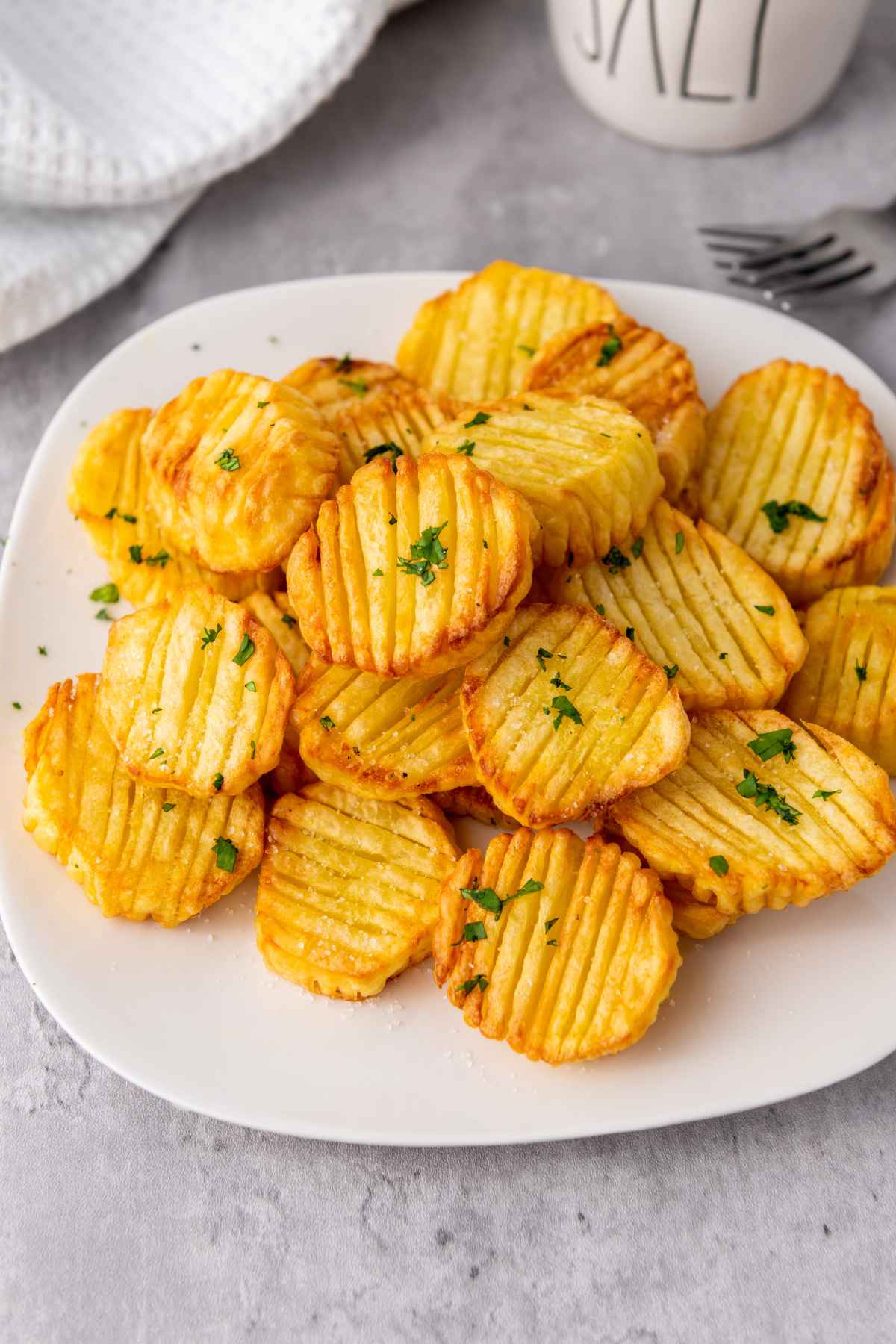 Angled view of a plate of golden brown accordion potatoes garnished with chopped parsley served on a white plate with a soft gray background.